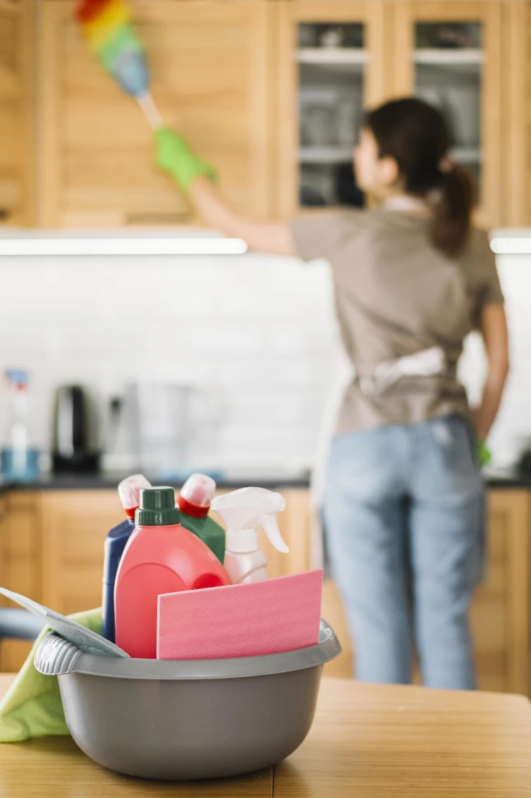 Cleaner dusting kitchen cabinets with cleaning supplies in foreground Cleaner dusting kitchen cabinets with cleaning supplies in foreground